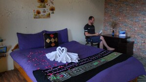 A room with a bed, a desk, and a chair. The bed has a purple blanket and pillows. There is a bouquet of flowers on the wall. - Sapa Tatu Homestay (Sa Pa)
