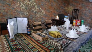 A dining table set with a menu, a bottle of beer, a basket of utensils, a plate of bananas, and a decorative tablecloth. - Sapa Tatu Homestay (Sa Pa)