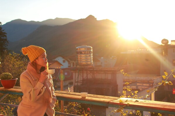 A person standing on a balcony with a cup, overlooking a mountainous landscape and buildings in the background.