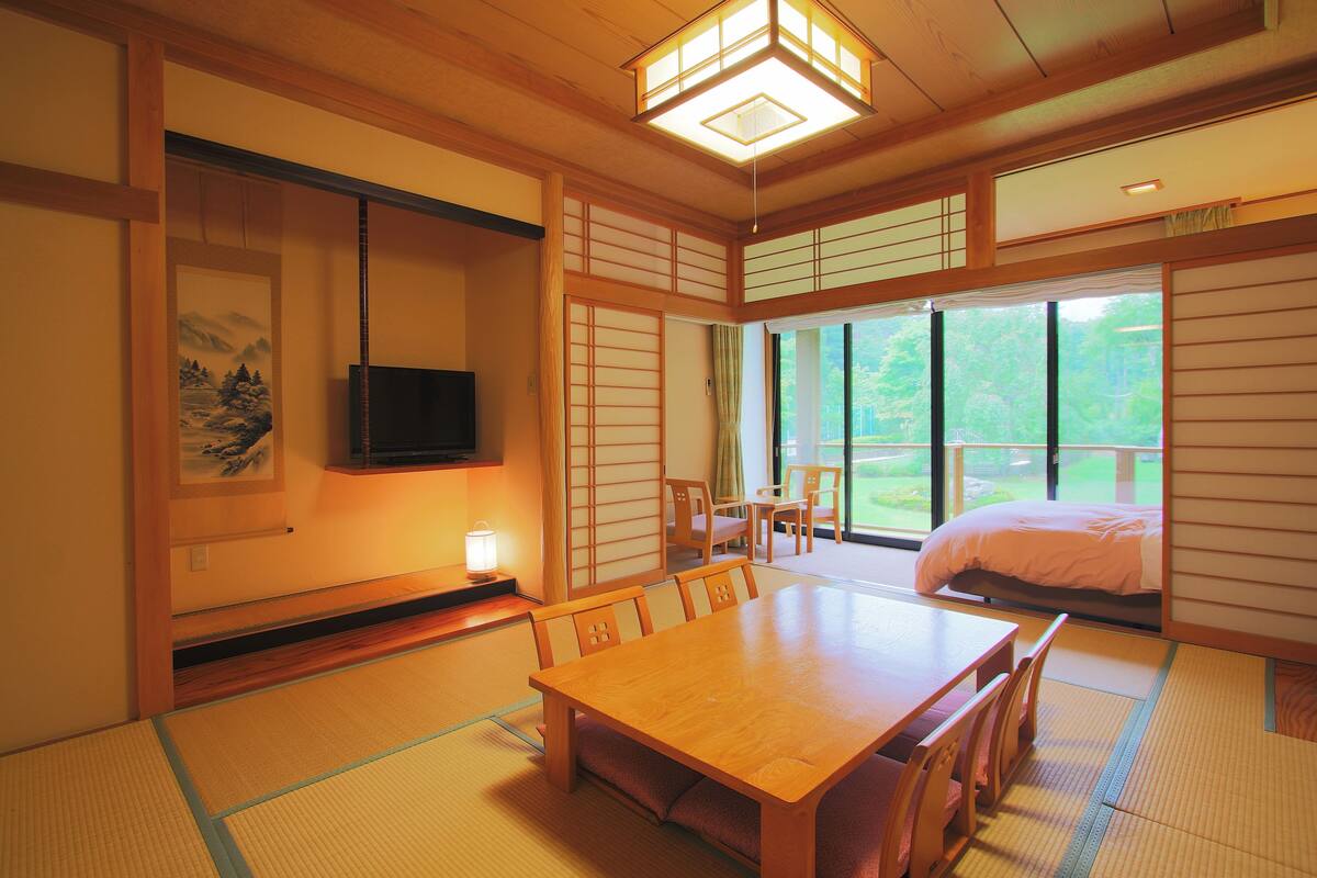 Perinteinen japanilainen huone, jossa on tatamimatto, puupöytä, tuoleja, televisio ja suuri ikkuna, josta avautuu näkymä vehreään maisemaan.