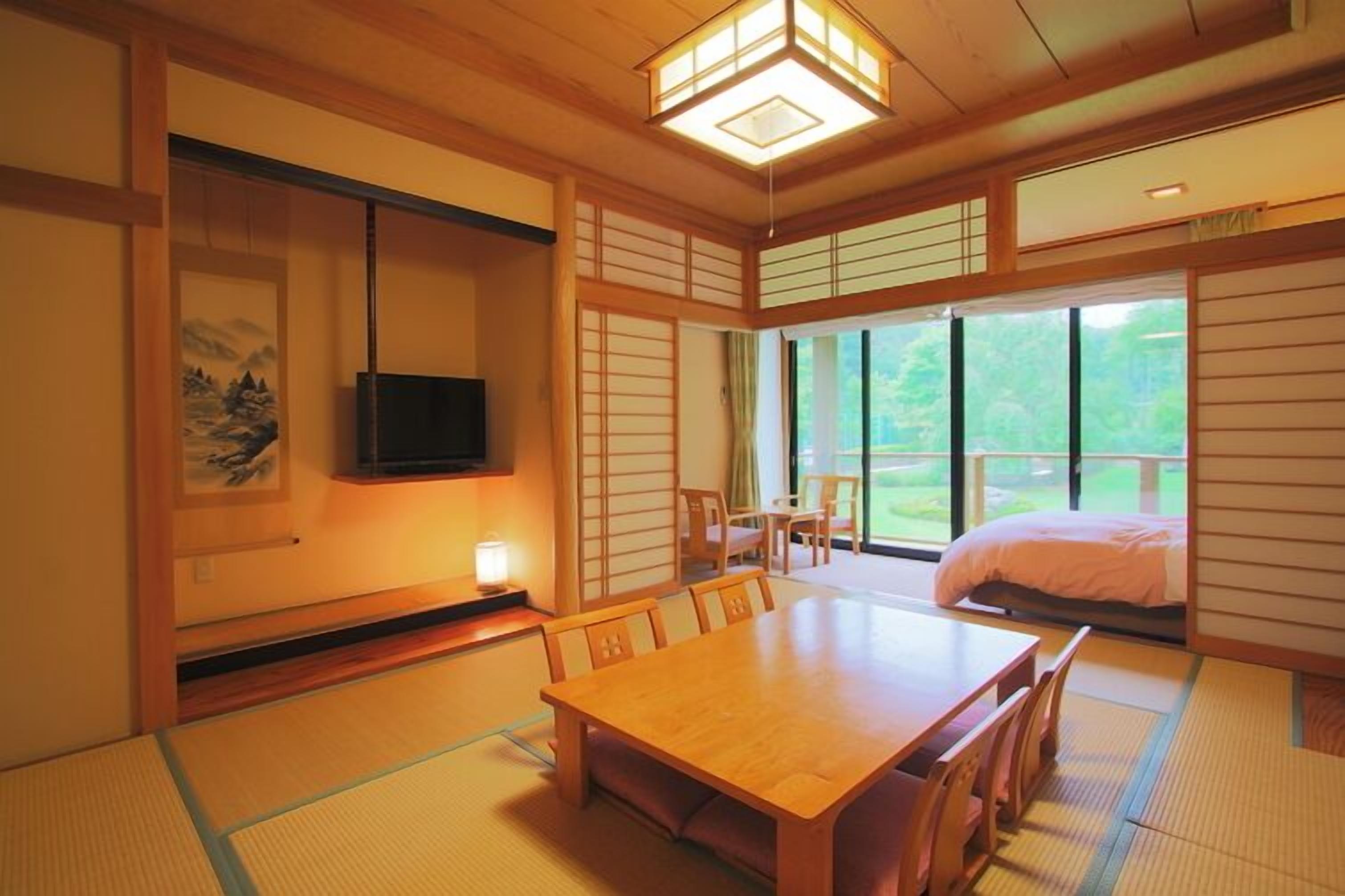 Um quarto japonês tradicional com tatames, uma mesa de madeira para refeições, uma televisão e uma cama com vista para áreas verdes.