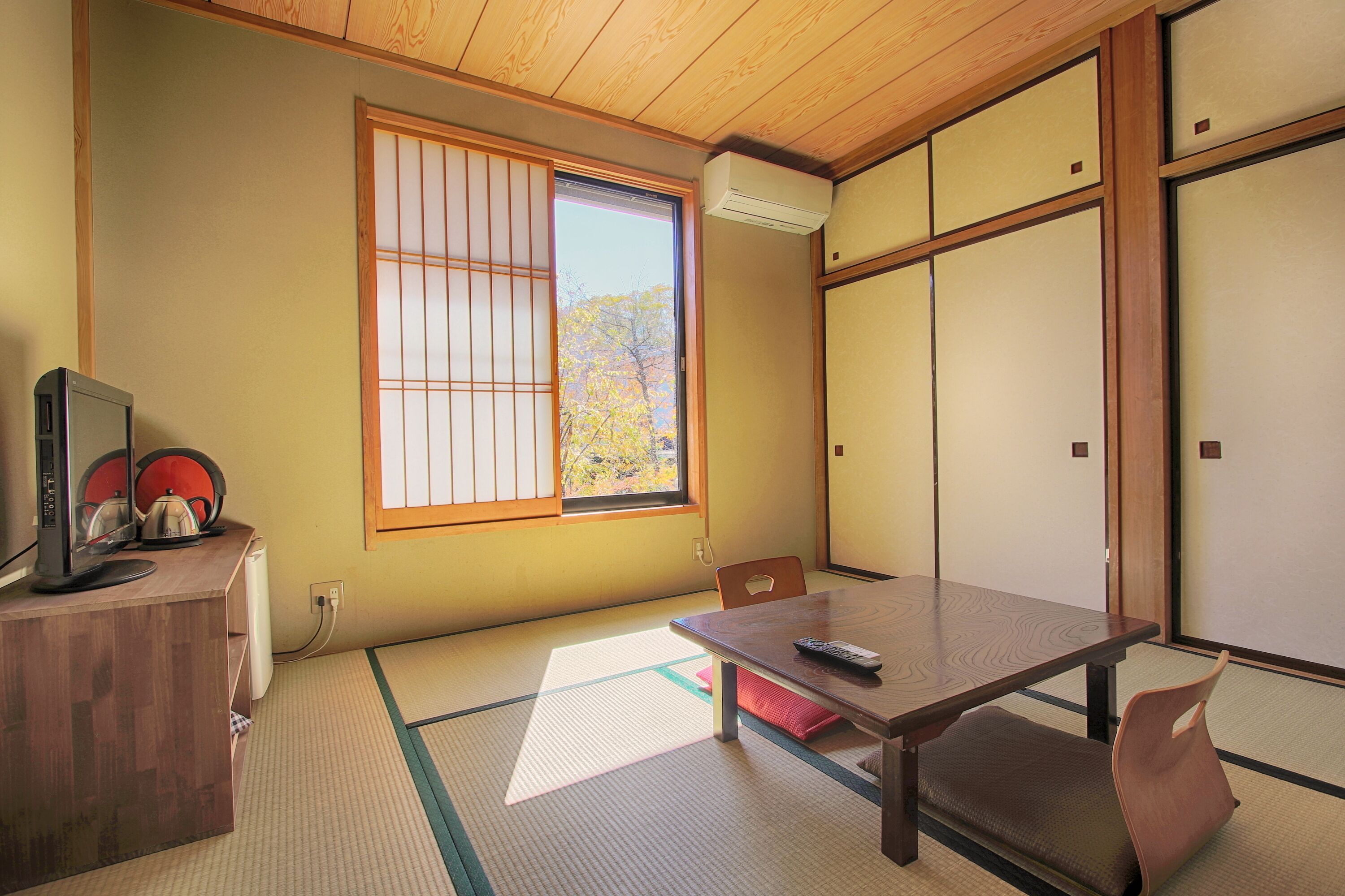 A traditional Japanese room with tatami flooring, a wooden desk with a computer, a table with a remote control, and sliding doors.