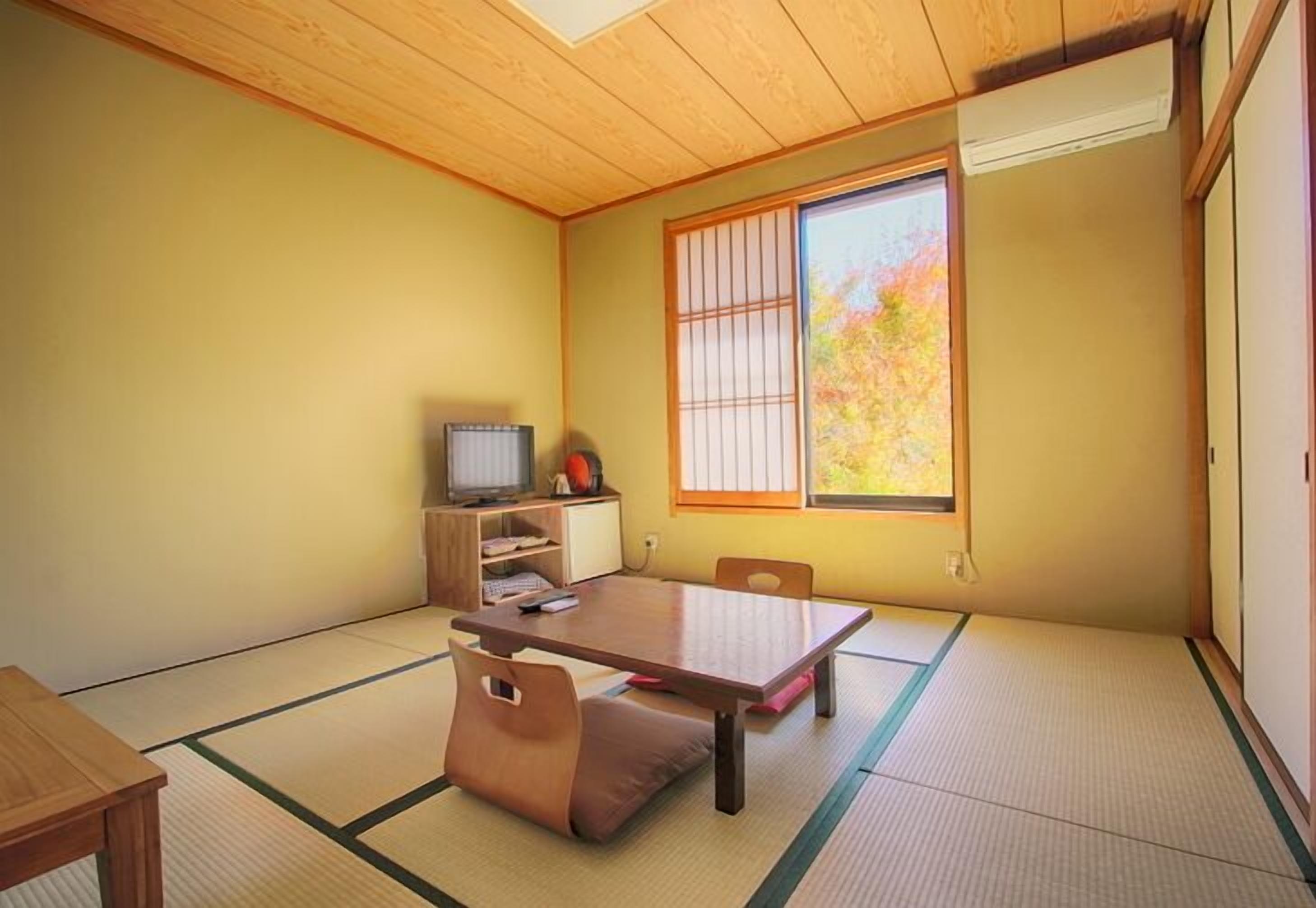 A traditional Japanese room with tatami flooring, a low table, a television, and a window with a view of autumn foliage.