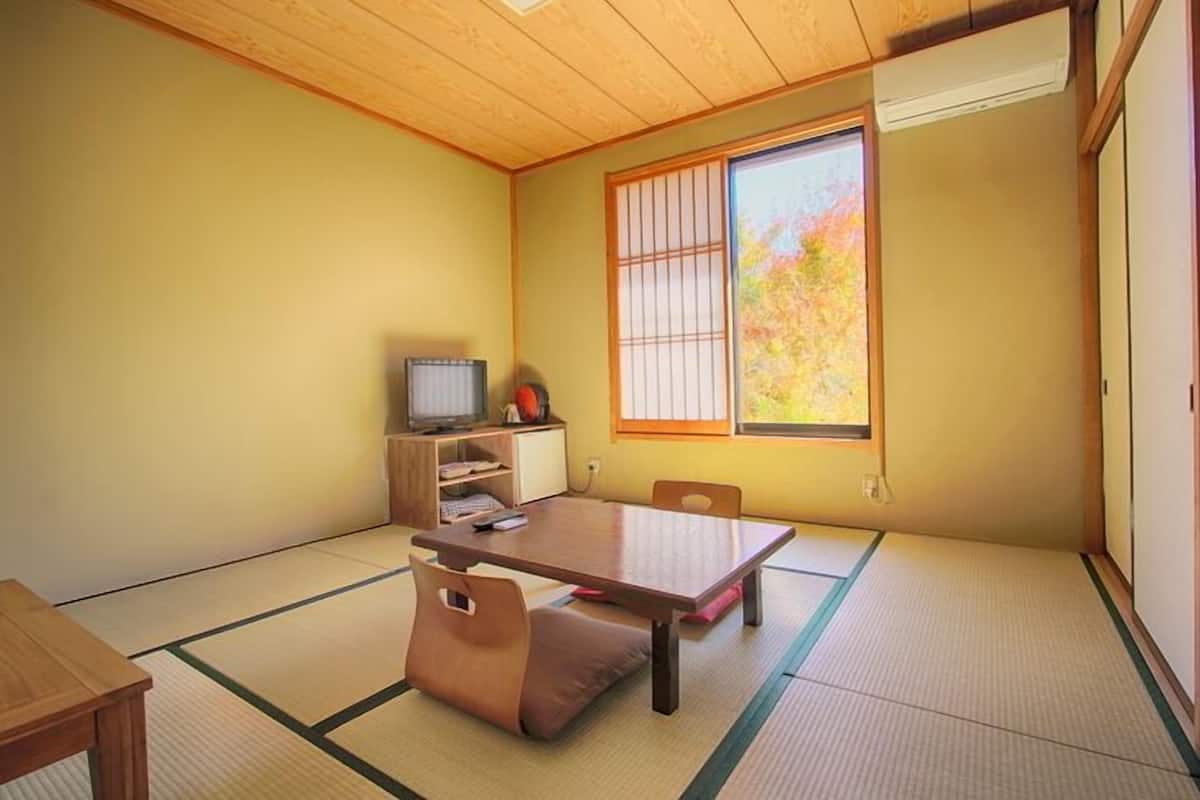 A traditional Japanese room with tatami flooring, a low table, a television, and a window with a view of autumn foliage.
