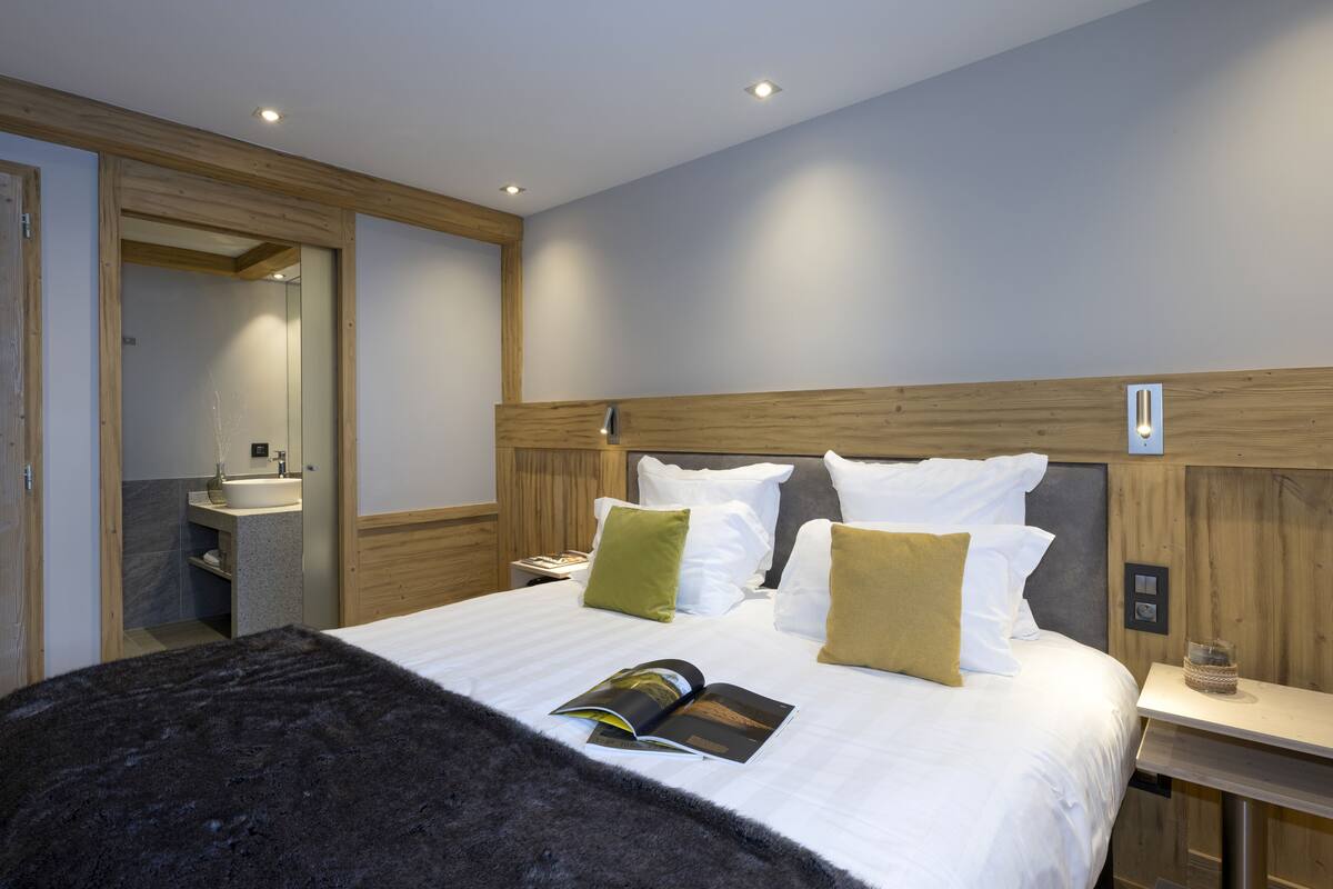 Una habitación de hotel moderna con una cama grande, cabecera de madera, una mesita de noche y un baño pequeño con lavamanos.