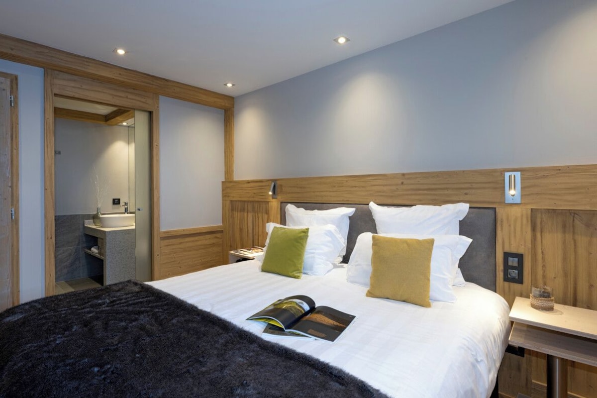 Una habitación de hotel moderna con una cama grande, cabecera de madera, una mesita de noche y un baño pequeño con lavamanos.