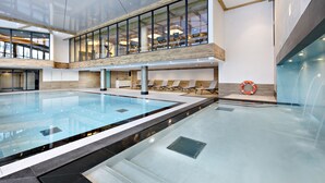 Una piscina interior moderna con paredes de vidrio transparente, sillones de descanso y estructura de techo de madera.