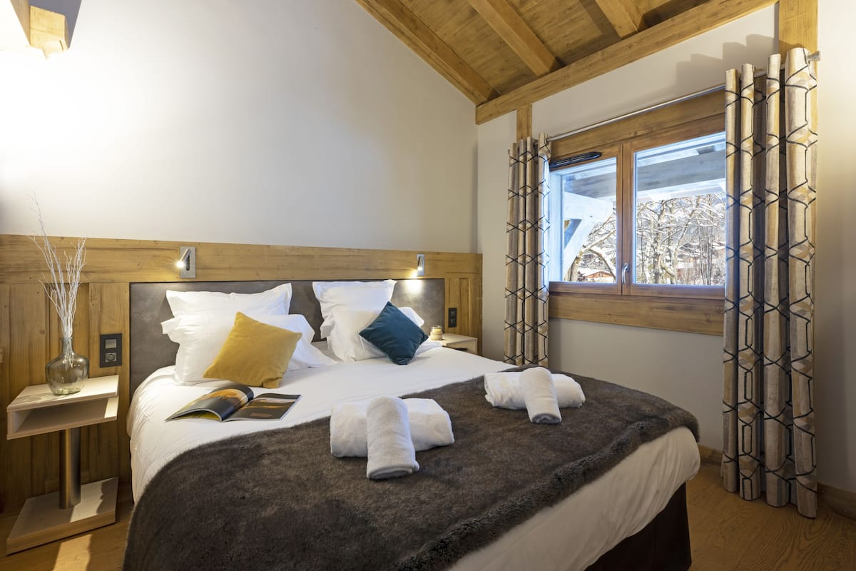 Una cama bien hecha con almohadas, una manta y toallas, una mesita de noche con un florero, y una ventana con cortinas.