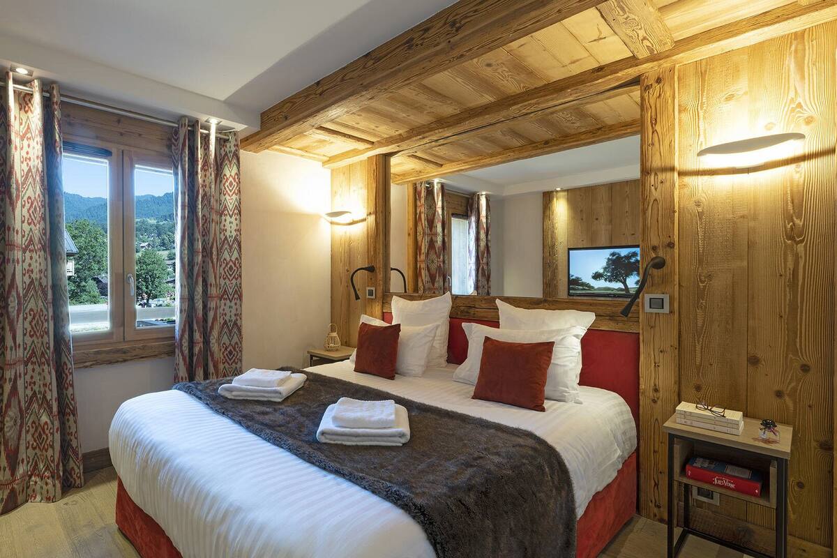 A bedroom with a wooden ceiling, a large bed with white and red bedding, a bedside table with a lamp, and a view of the outdoors through a window.