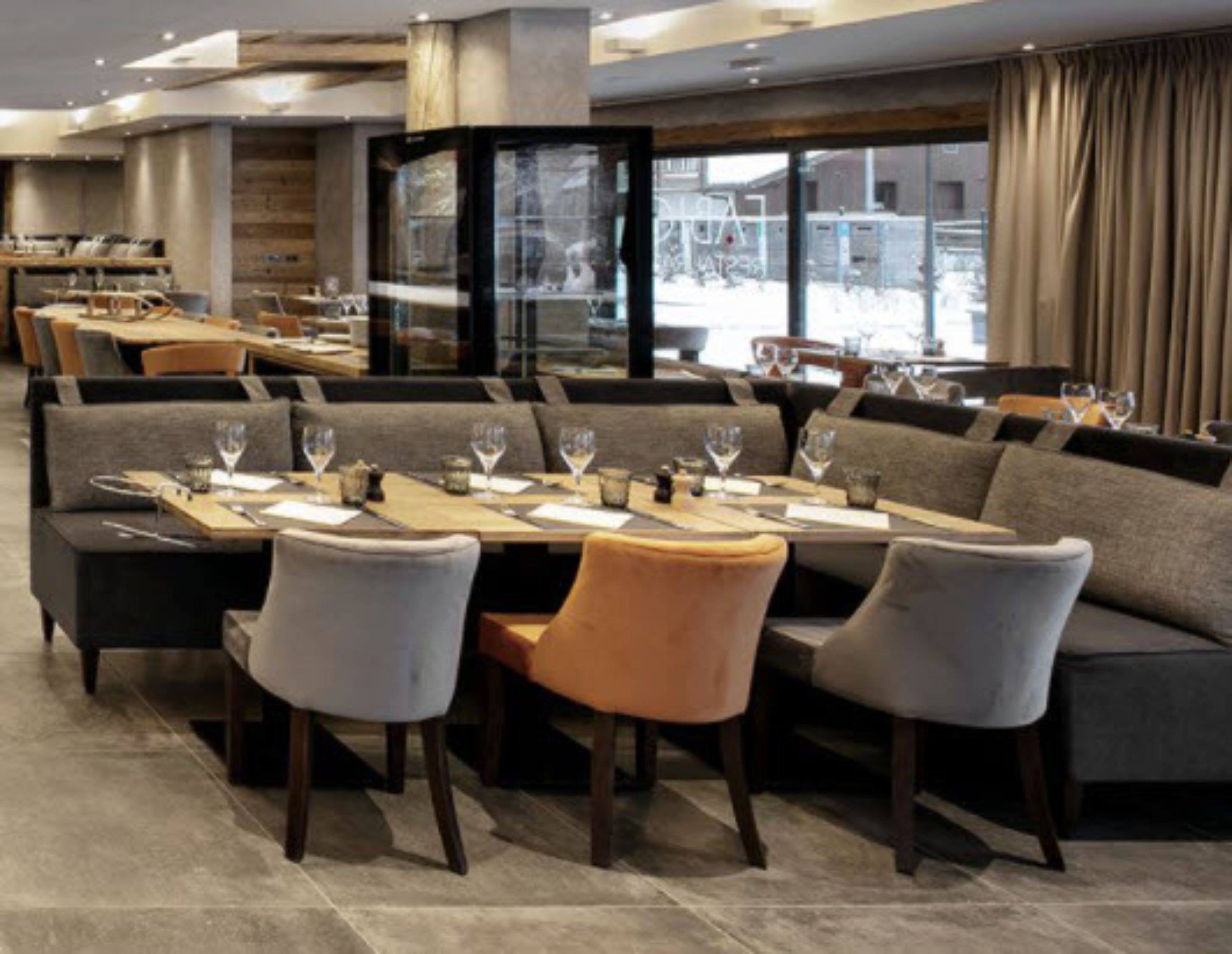 A modern dining area with a long table set for a meal, cushioned seating, and a large window offering a view of the outside.