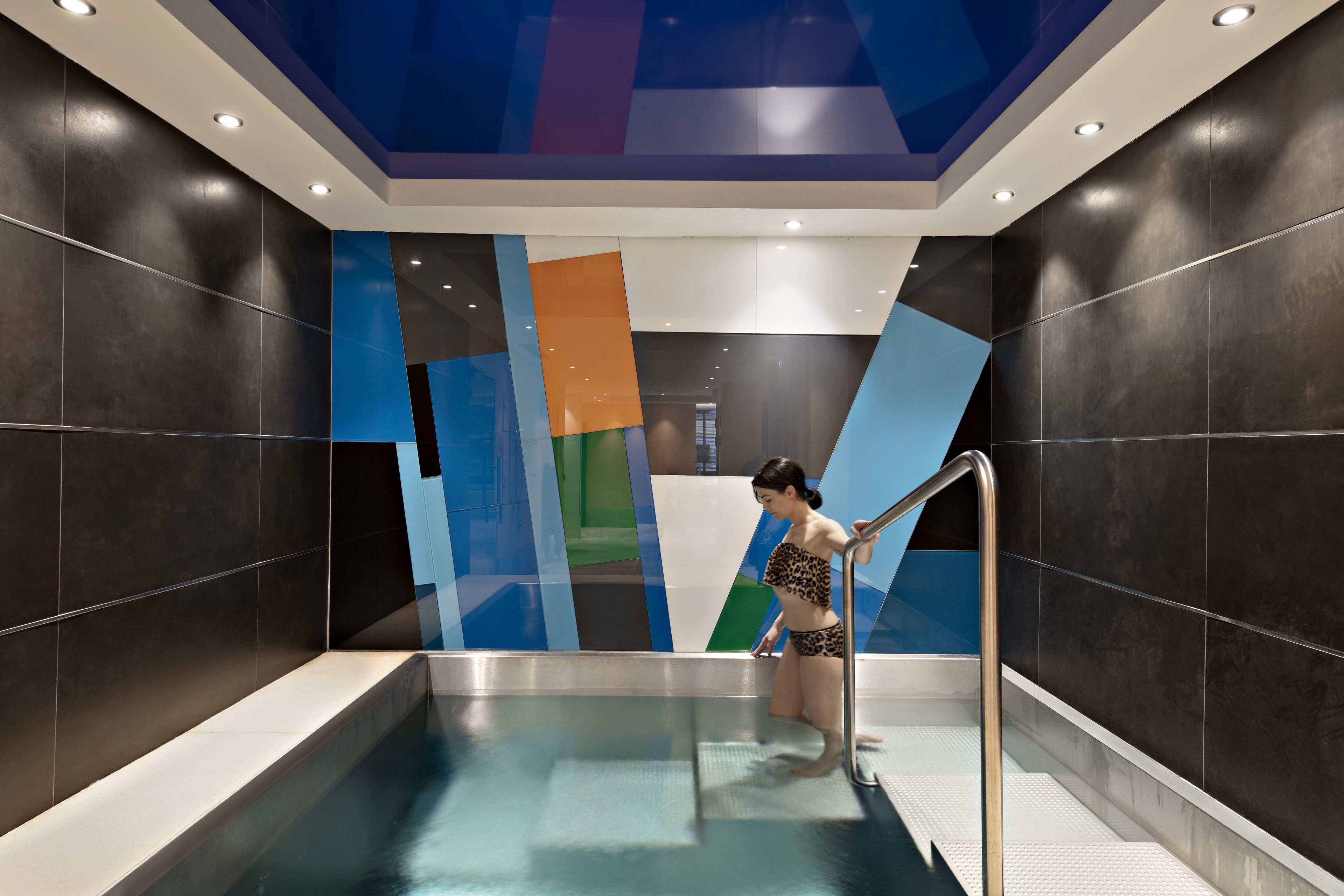 A modern indoor swimming pool with a geometric wall design and a person using the pool's ladder.