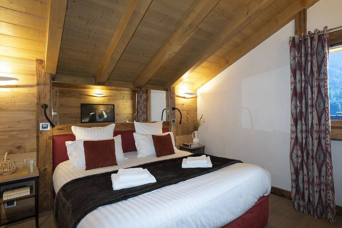 A cozy bedroom with a wooden ceiling, a large bed with white and red bedding, a bedside table with a lamp, and a window with curtains.