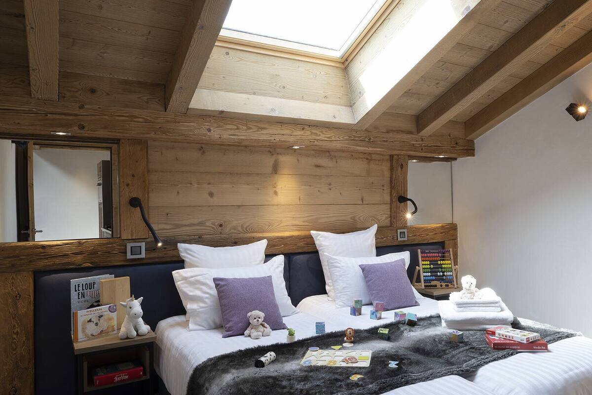 A bedroom with a sloped ceiling, wooden beams, a skylight, a bed with pillows, a nightstand with a teddy bear, and a wall-mounted lamp.
