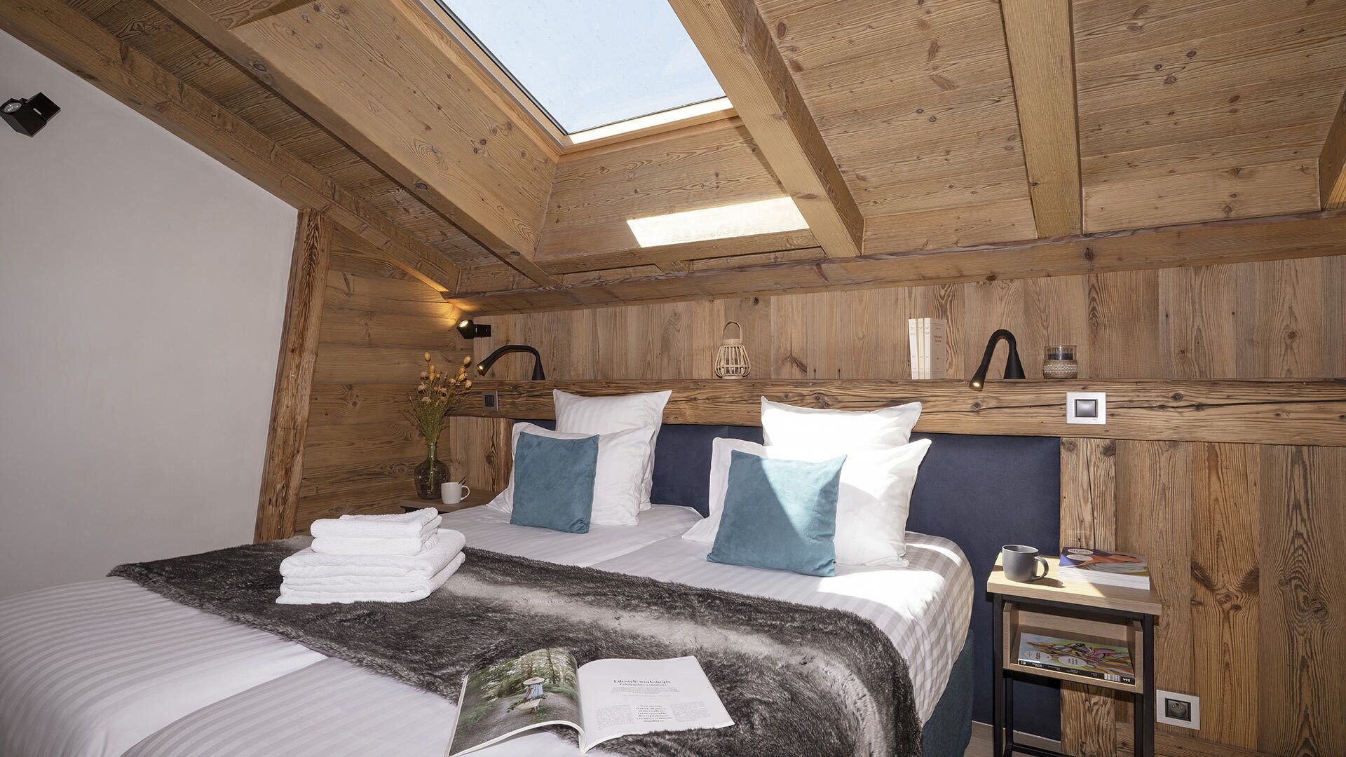 Un dormitorio acogedor con tragaluces, paredes de madera, una cama con ropa de cama blanca y azul, una mesita de noche con lámpara y una pequeña mesita auxiliar.
