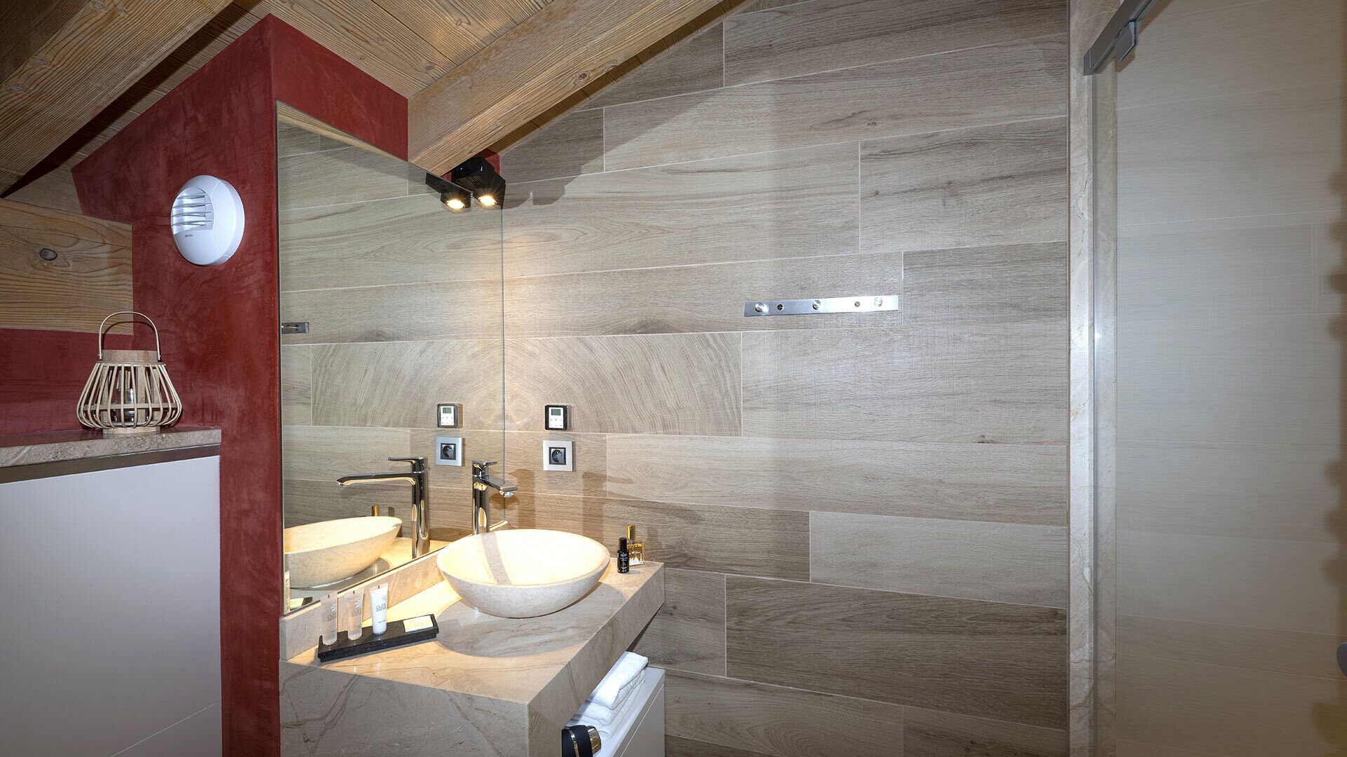 A modern bathroom with a large stone sink, two faucets, and a mirror. There are wooden accents on the ceiling and a red accent wall.