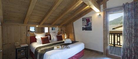 A bedroom with a wooden ceiling, a bed with white and red bedding, a nightstand, and a view of a mountain through a window.
