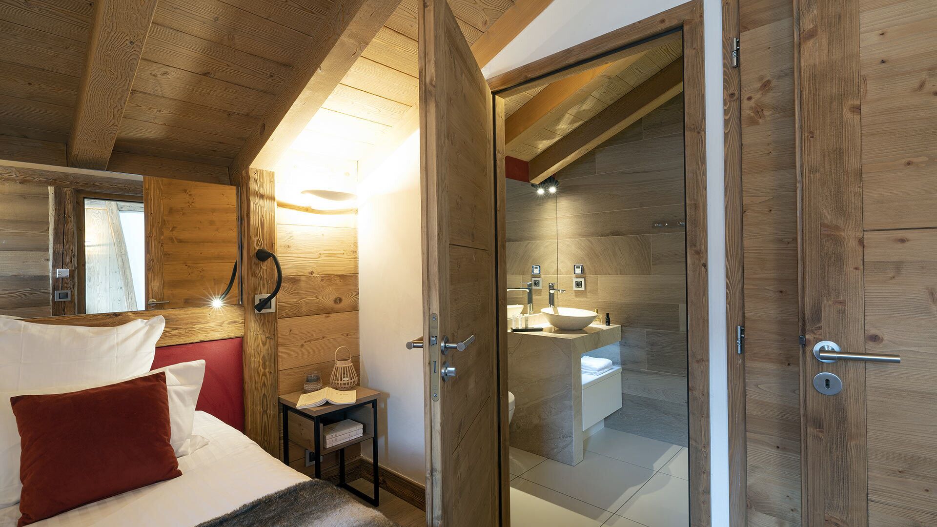 A bedroom with a wooden ceiling, a bed with white and red bedding, a bedside table with a lamp, and a small nightstand with books.