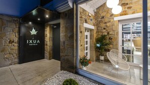 Exterior del HOTEL IXUA con fachada de piedra, una puerta de entrada negra y un área de estar visible a través de las puertas de vidrio.