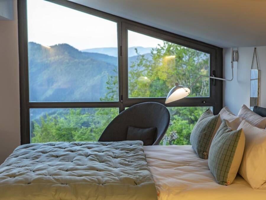 Quarto com uma janela grande, cama com roupa de cama branca, uma cadeira e uma luminária de mesa.