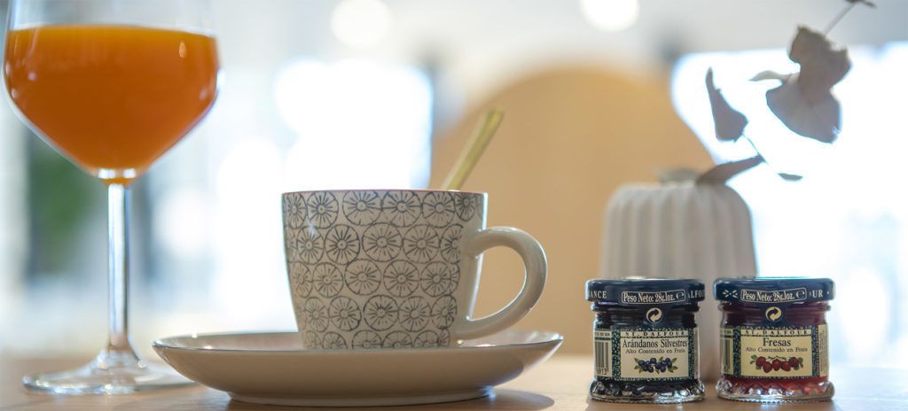 A table with a patterned cup, a glass of orange juice, and two jars of preserves.
