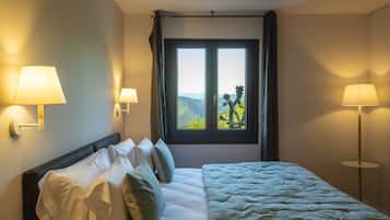 Ein Hotelzimmer mit Bett, zwei Nachttischlampen, einem Fenster mit Vorhängen und Blick auf Grünfläche.