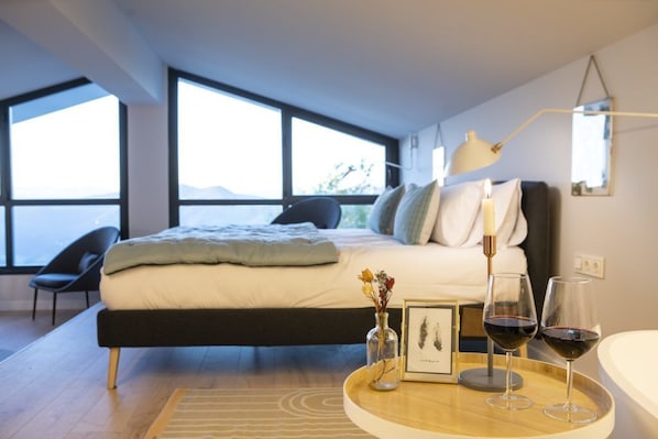 Un dormitorio moderno con una cama grande, una mesita de noche con un jarrón y un marco de fotos, y dos copas de vino tinto.