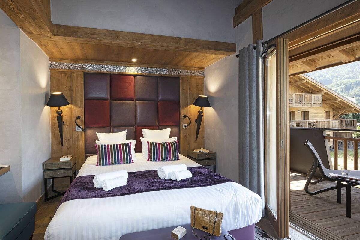Un dormitorio con una cama grande, cabecera de madera, dos mesitas de noche con lámparas, un banco y vistas a una terraza de madera y montañas.