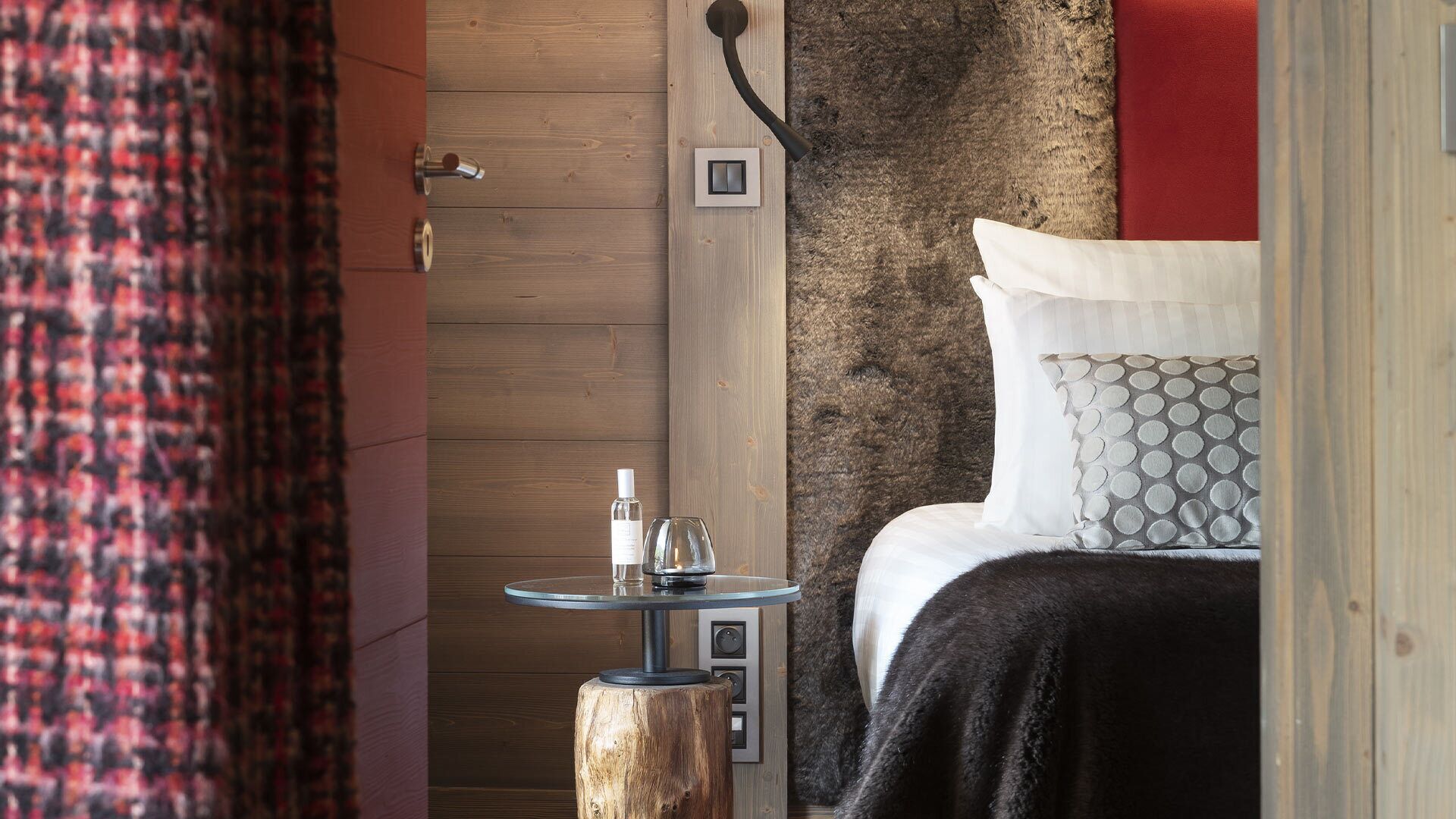 Un dormitorio acogedor con puerta de madera, una mesita de noche pequeña con una botella y un vaso, y una cama con almohadones estampados.