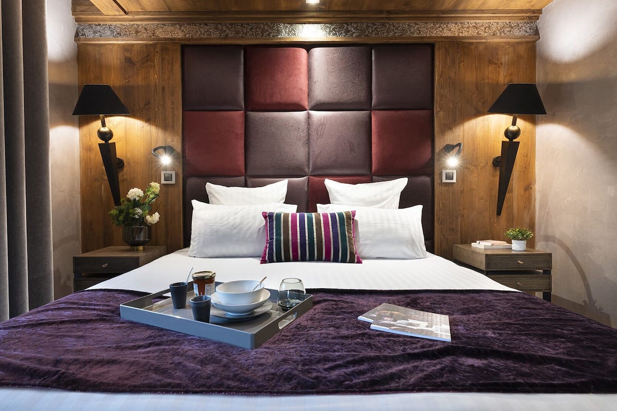 Habitación de hotel con una cama grande, cabecera de madera, dos mesitas de noche con lámparas, una manta morada y una bandeja con alimentos para el desayuno.