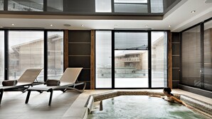 Una piscina interior moderna con paredes de vidrio transparente, sillones de descanso y vistas a edificios exteriores.