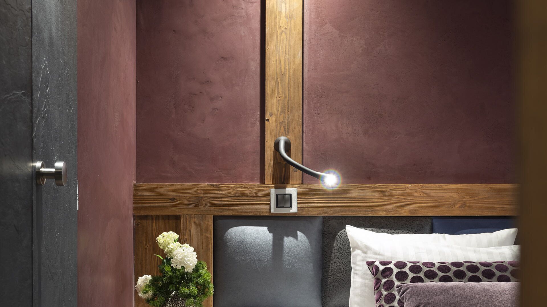 A room with a wooden door, a wooden ceiling, a light fixture, a bed with pillows, and a small plant.