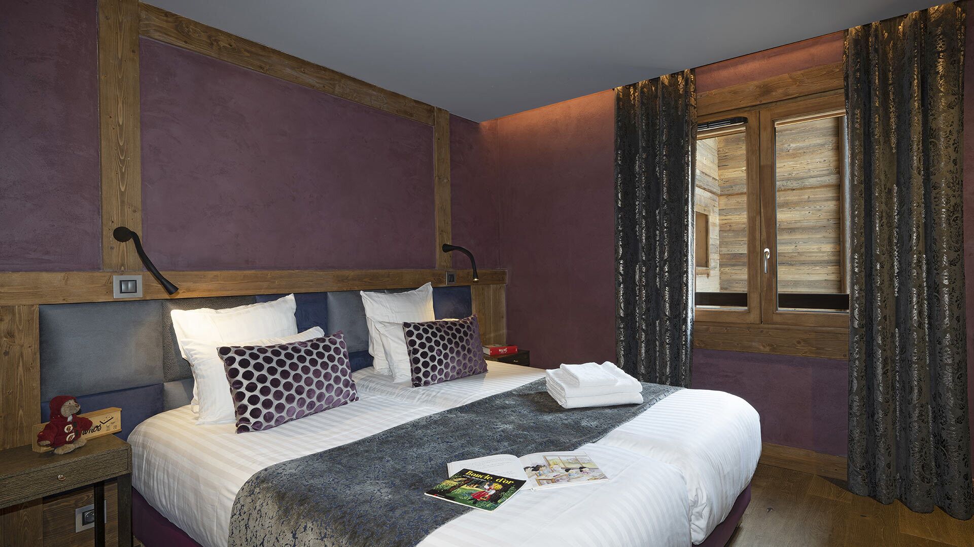 A hotel room with a large bed, wooden headboard, purple walls, a window with curtains, and a nightstand with a teddy bear.