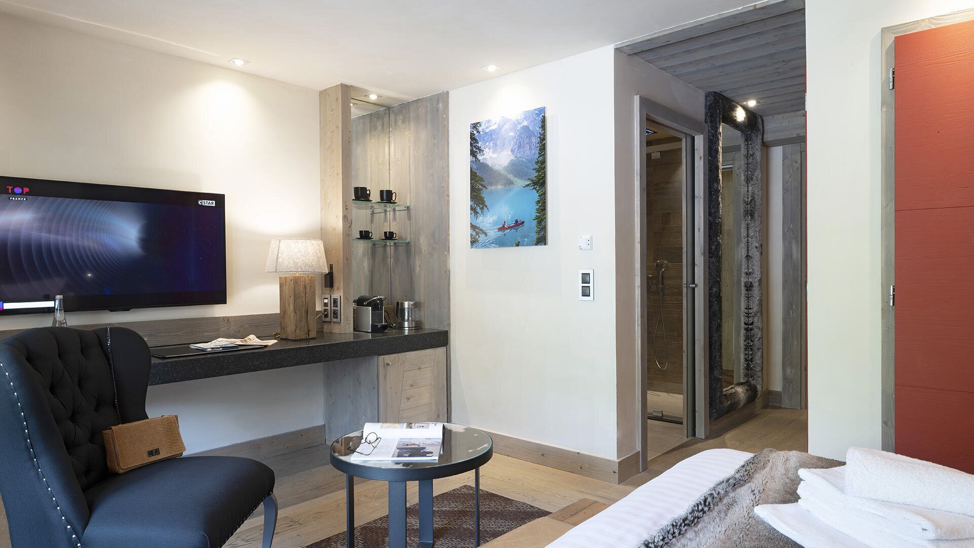 A hotel room with a flat-screen TV, a sofa, a small table, a painting, a shower, and a bed with towels.