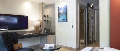 A hotel room with a flat-screen TV, a sofa, a small table, a painting, a shower, and a bed with towels.