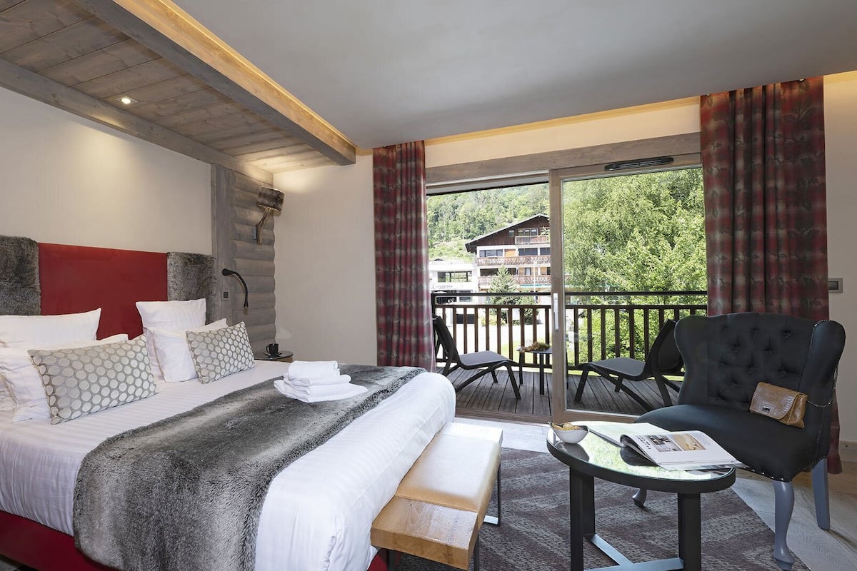 Una habitación de hotel con una cama grande, cabecero de madera, balcón con vistas al exterior y una silla.