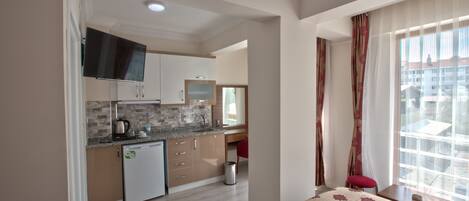 Habitación de hotel compacta con kitchenette, cama con colcha estampada, ventana con cortinas translúcidas y vistas a edificios.