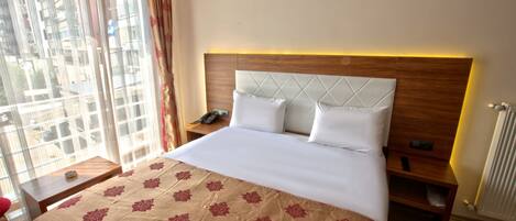 A hotel room with a large bed, a wooden headboard, a bedside table with a phone, and a view of buildings through the window.