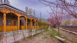 A row of traditional wooden buildings with a covered walkway, surrounded by trees and a mountainous backdrop. - Shangrila Resort Nubra (Leh)