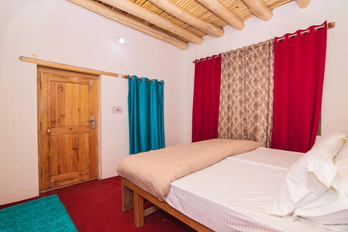 A bedroom with a wooden door, a bed with white bedding, and red and patterned curtains.