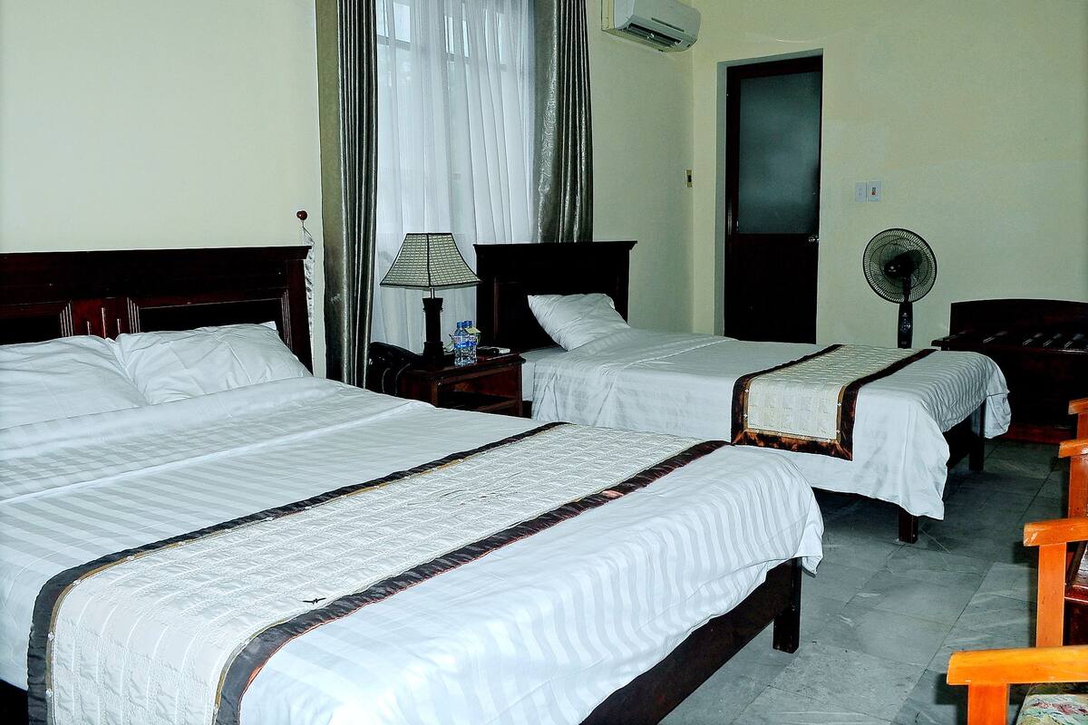 Une chambre d’hôtel avec deux lits, une table de chevet avec une lampe, un climatiseur et un ventilateur sur pied.