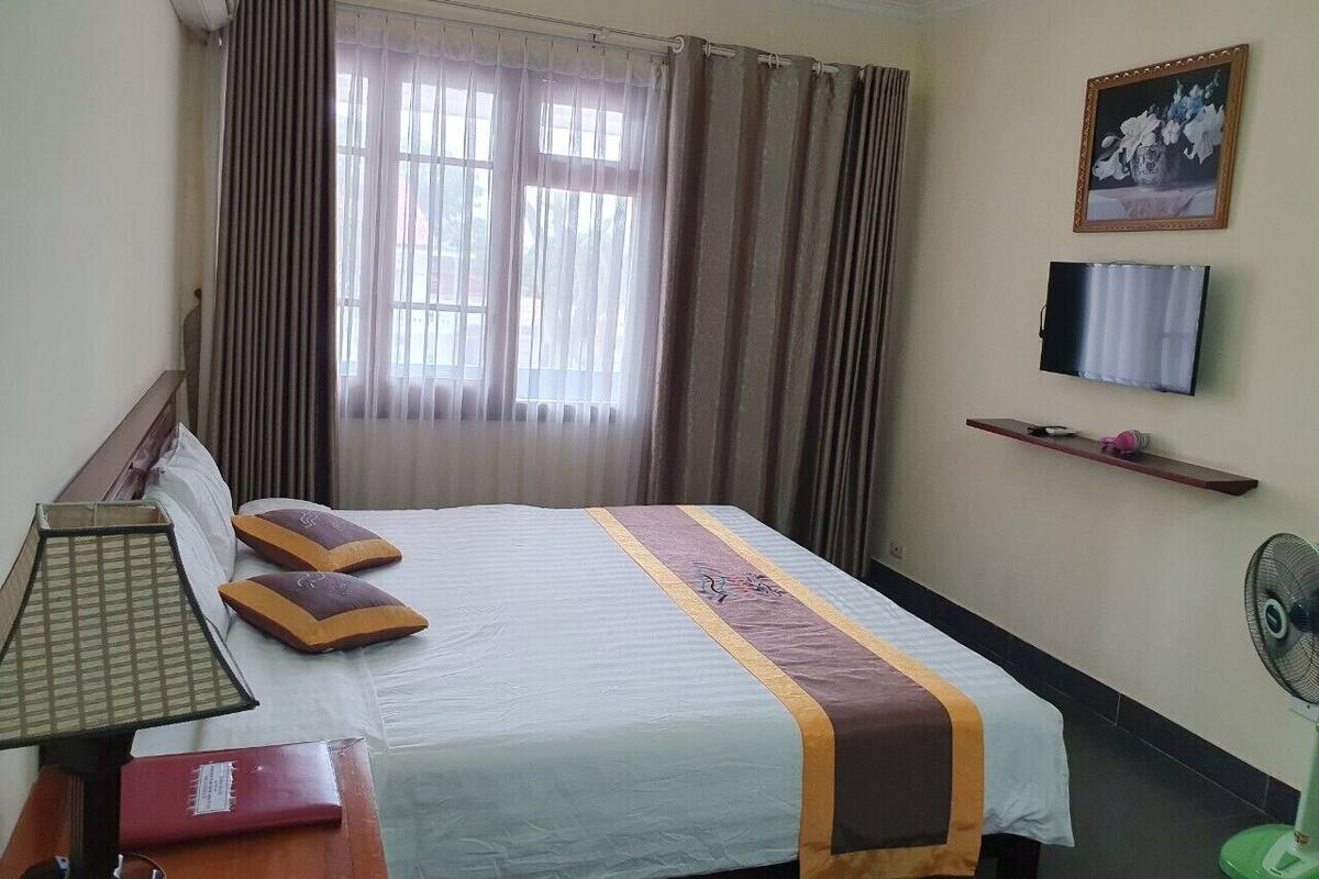 Une chambre d’hôtel avec un lit, un bureau avec une lampe, une télévision, un ventilateur et une fenêtre avec des rideaux.