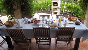 Un espace de restauration en plein air avec une table dressée pour le petit-déjeuner, entourée de chaises, et un four en pierre en arrière-plan.