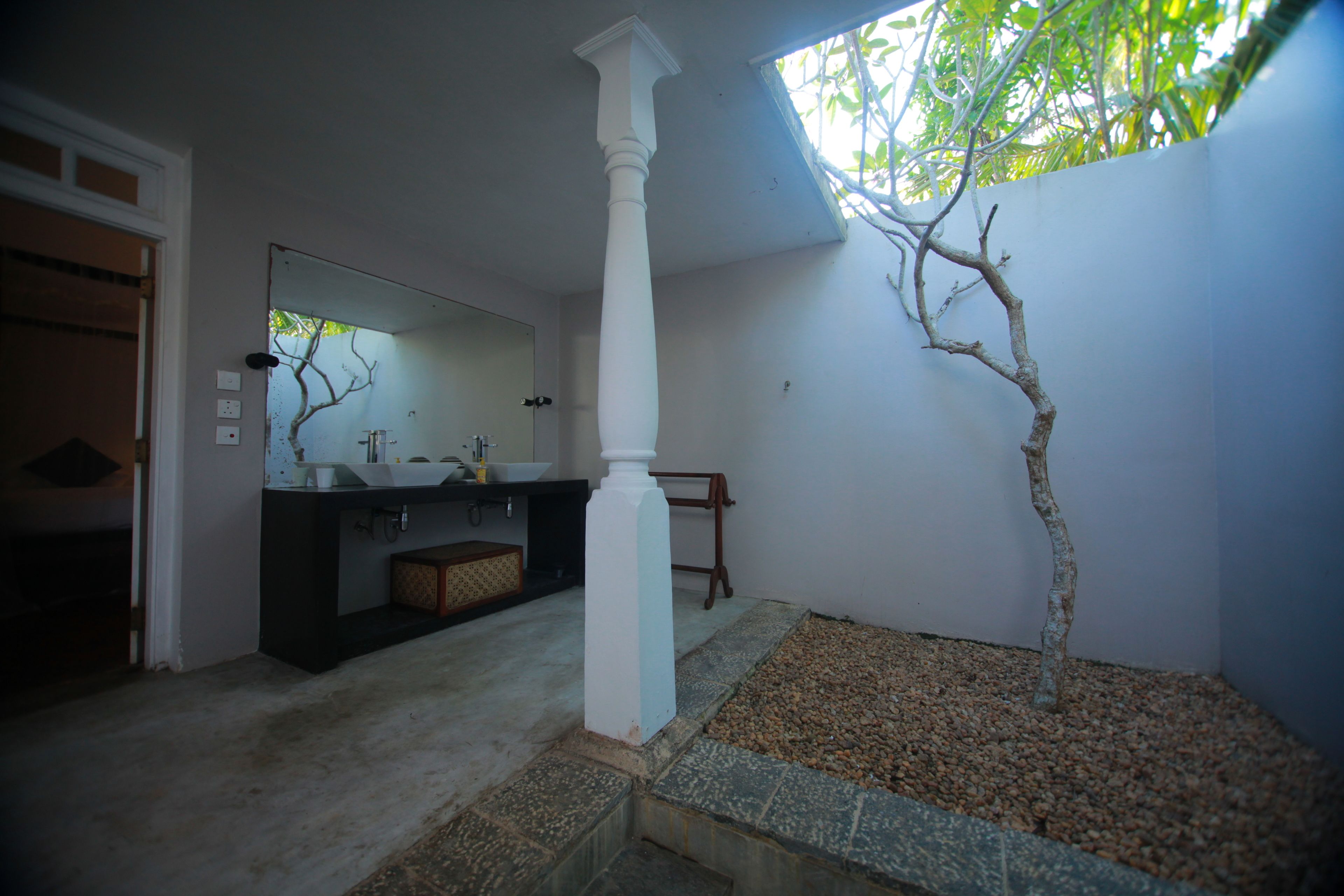 A bathroom with a white pillar, a sink, and a mirror. There is a tree in a planter and a wooden bench.