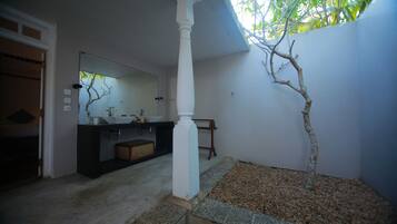 A bathroom with a white pillar, a sink, and a mirror. There is a tree in a planter and a wooden bench.