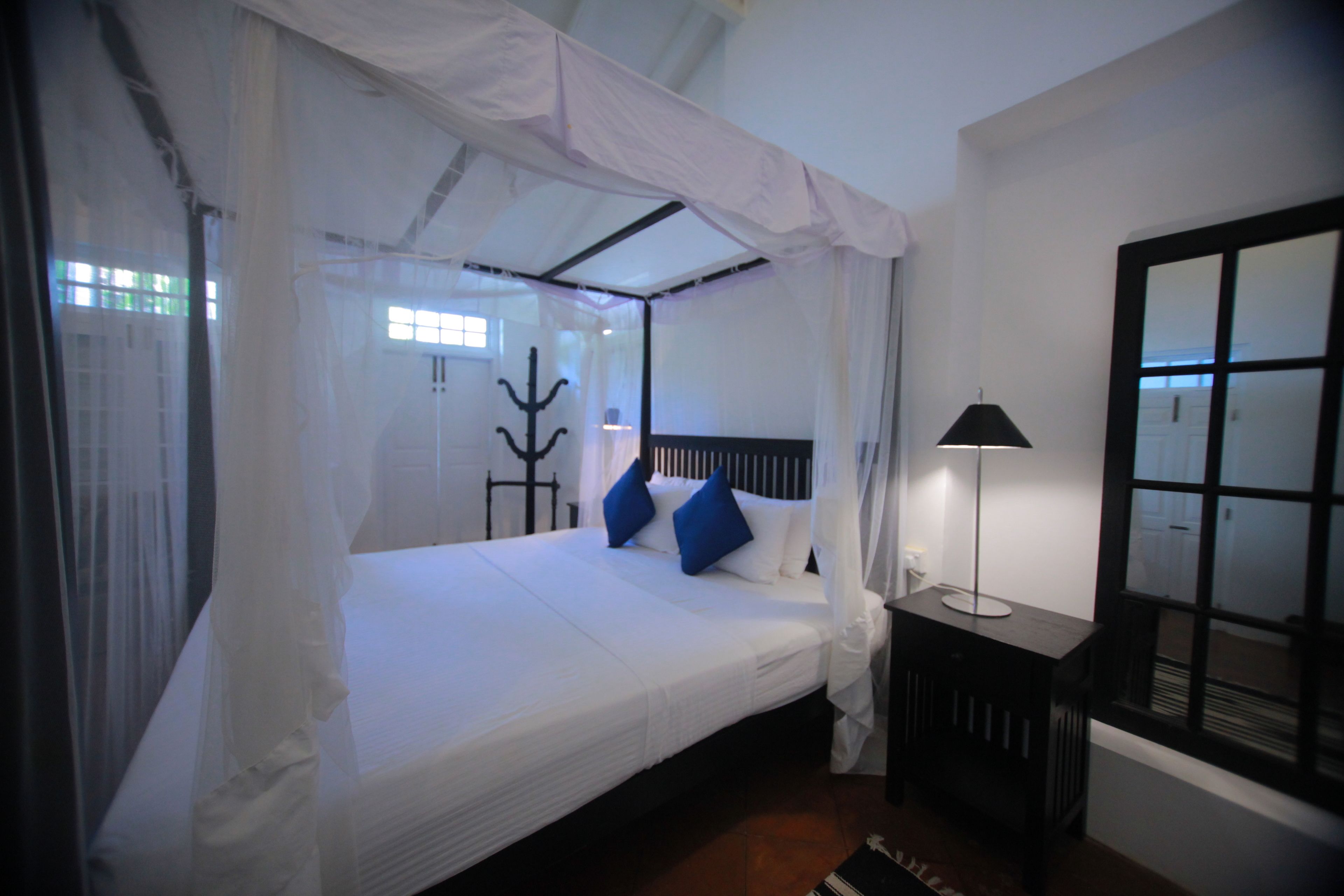 A bed with a canopy, a bedside table with a lamp, and a window with blinds.