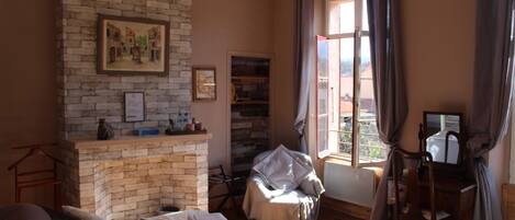 A room with a brick fireplace, a bed, a desk, and a window with curtains.