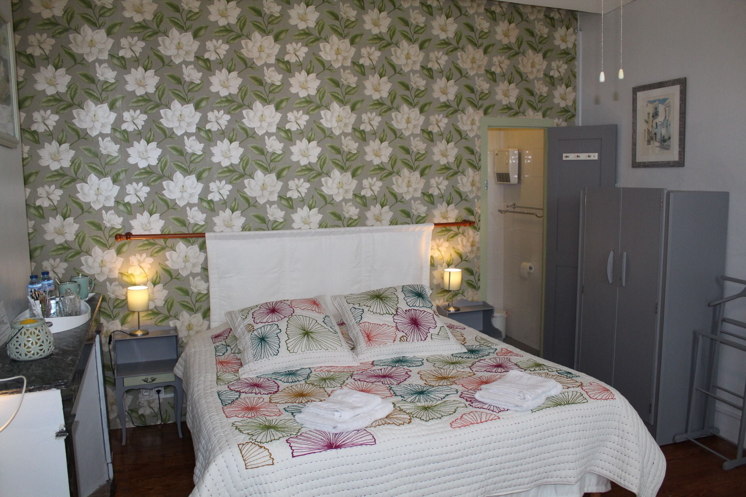 A bedroom with floral wallpaper, a bed with a quilt, bedside lamps, a wardrobe, and a small table with a lamp and decorative items.