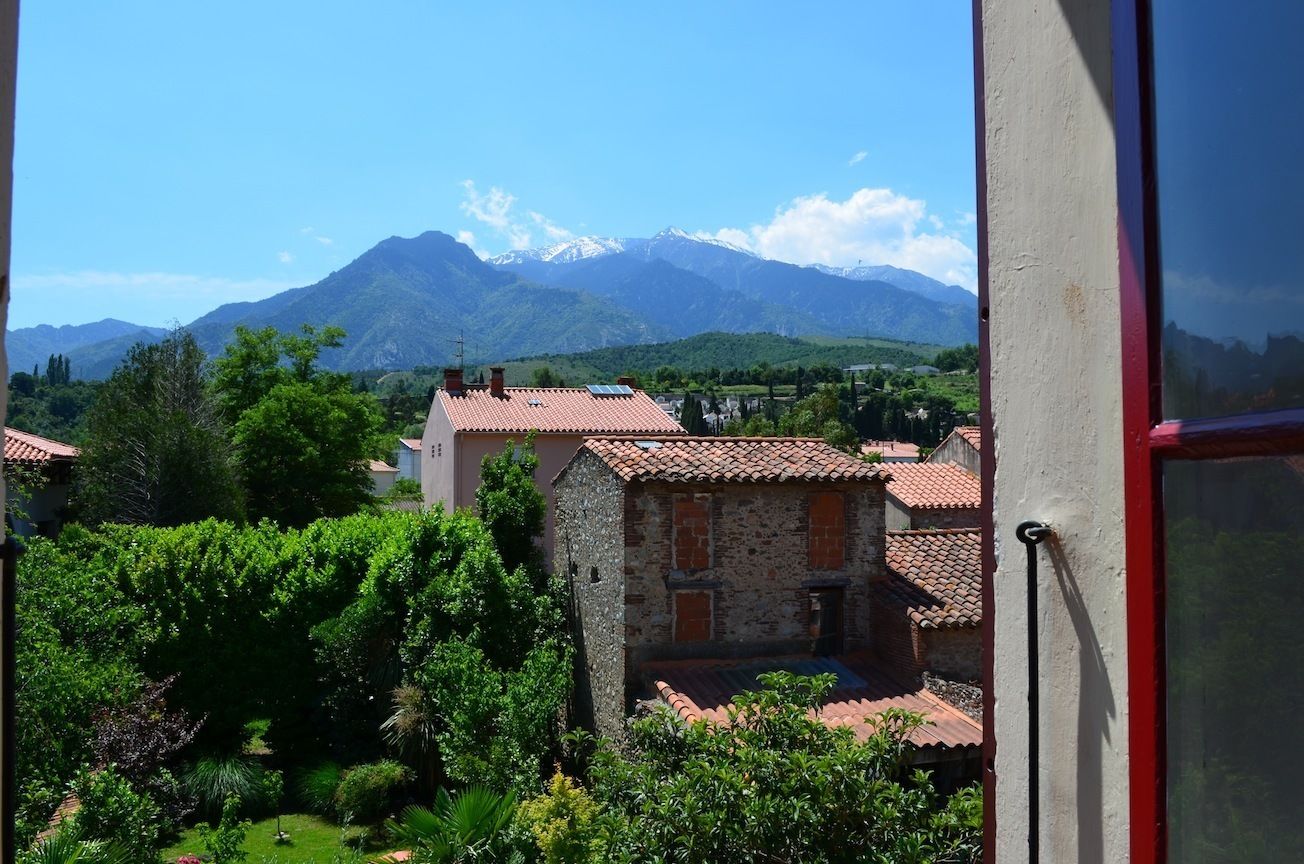 A view of a mountain range with snow-capped peaks in the background, a stone building with a red-tiled roof in the foreground, and lush green vegetation.