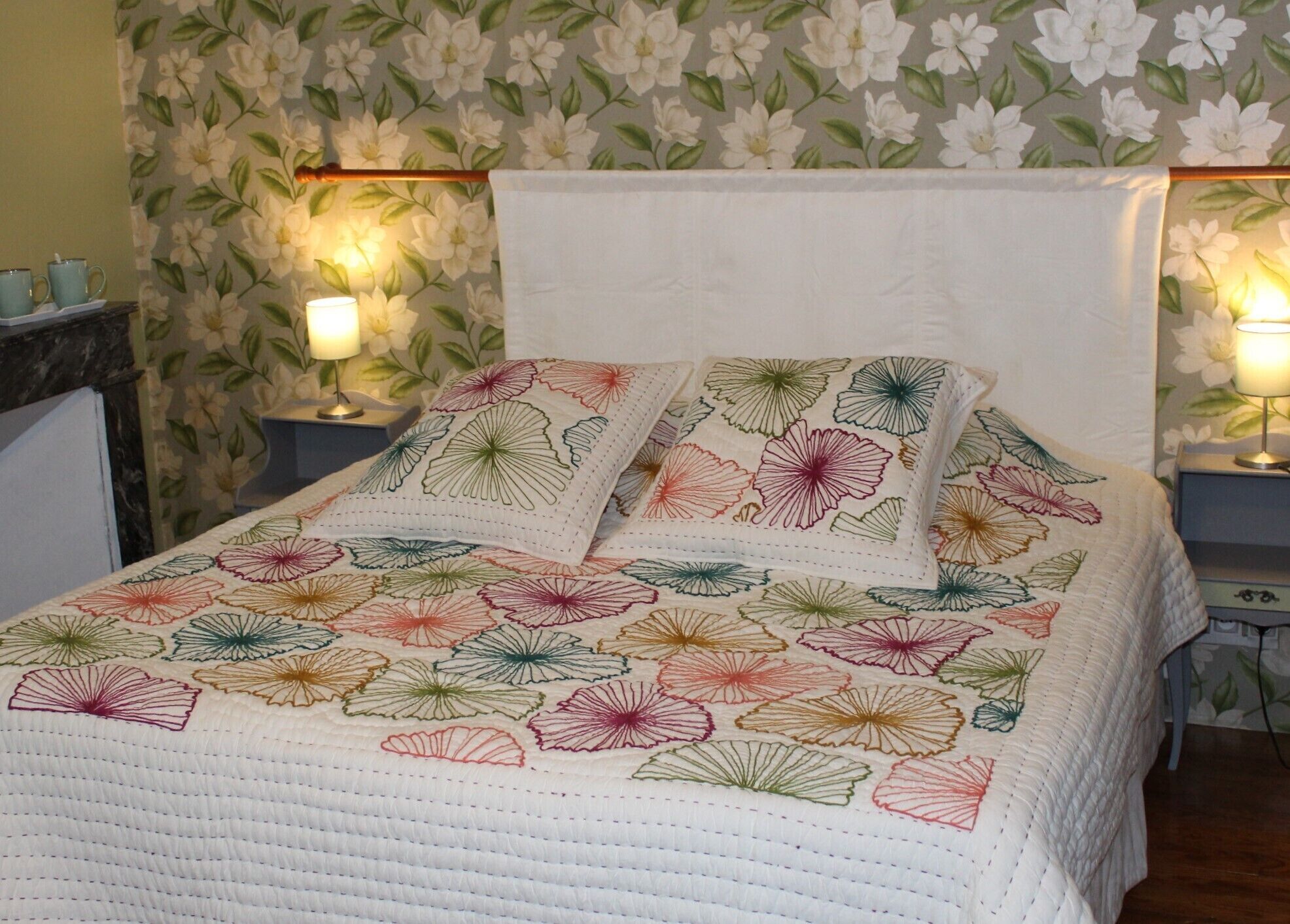 A bed with a floral-patterned quilt and pillows, two bedside lamps, and a floral wallpaper background.