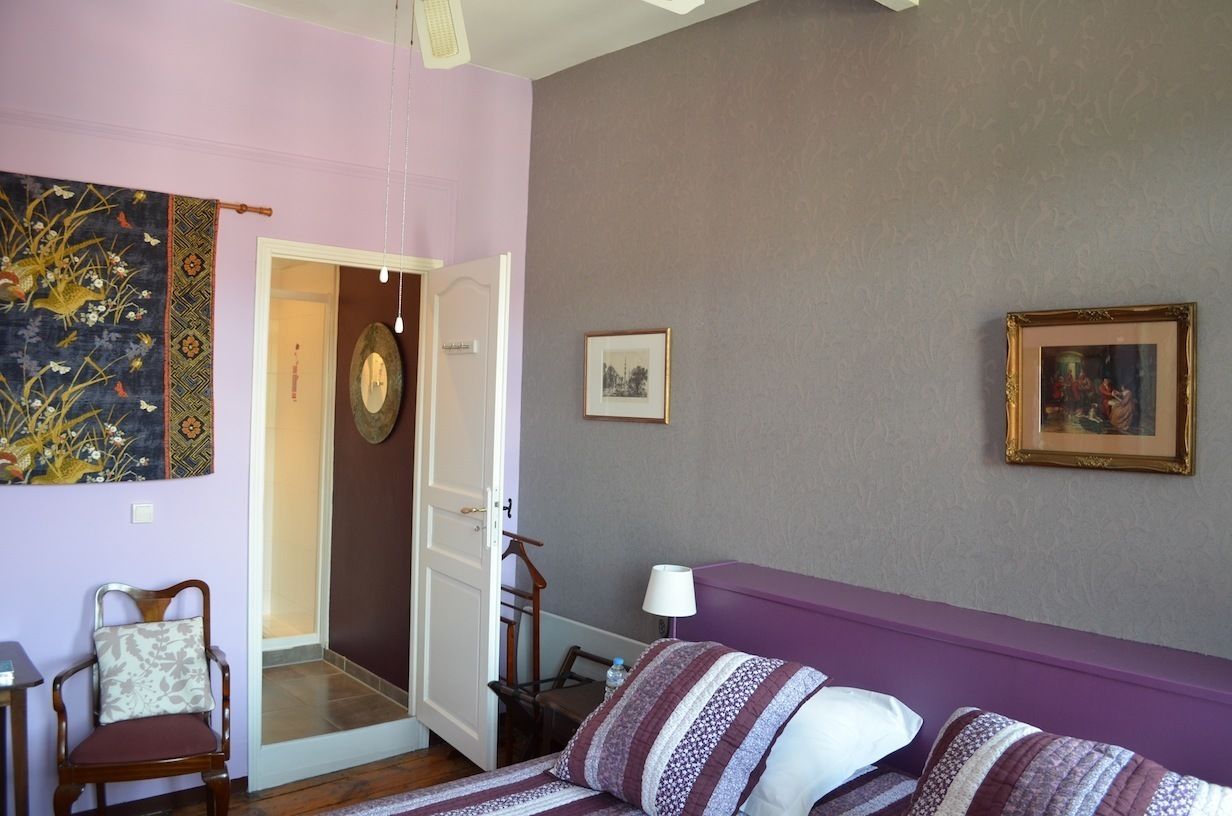 A room with a purple sofa, a wooden chair, a bed with striped bedding, a wall with a framed picture, and a door leading to another room.